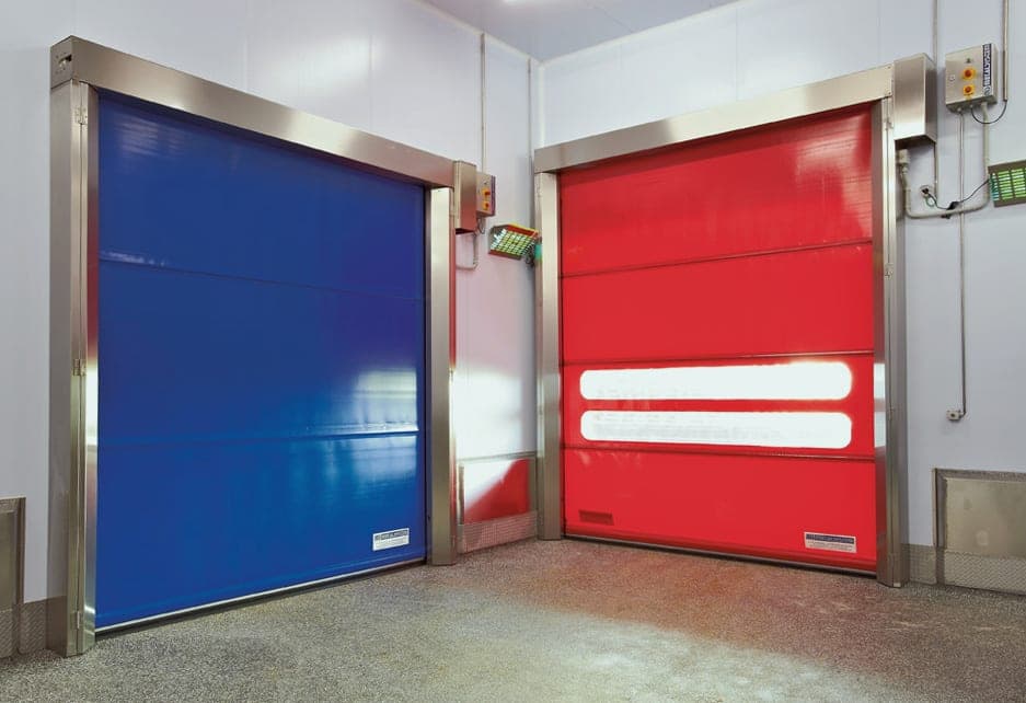 Dos puertas rápidas puestas lado a lado en un flujo de nave industrial. En la derecha se ve una cortina de color rojo con mirilla y en la izquierda una cortina de color azul.