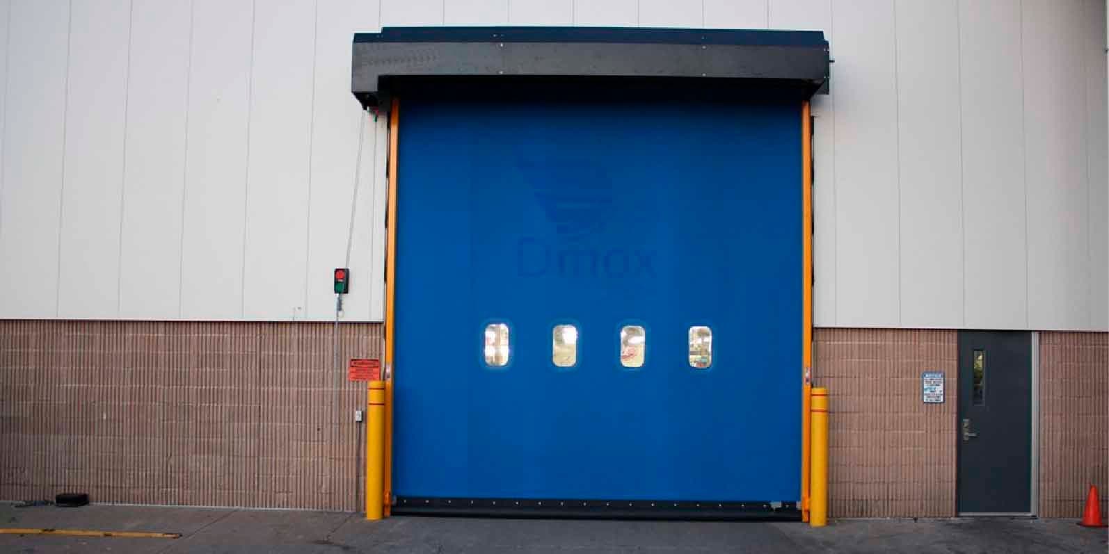 Puerta rápida azul de PVC con cuatro ventanillas de visión, instalada en un acceso exterior con capota y postes protectores