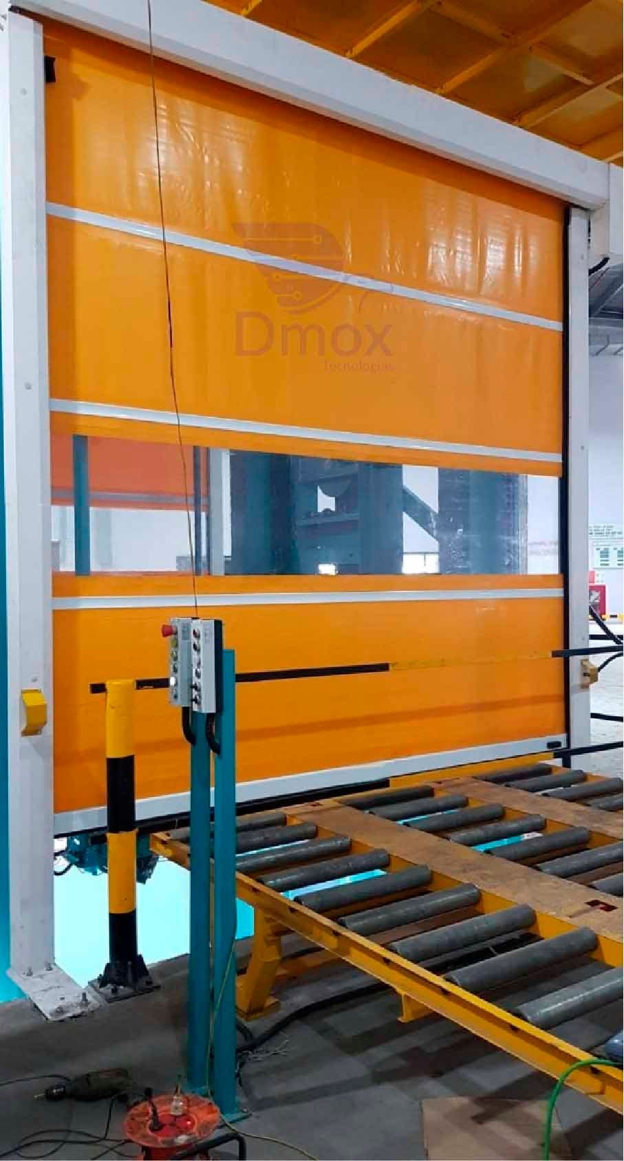 Puerta rápida naranja sobre una línea de rodillos, con poste protector amarillo