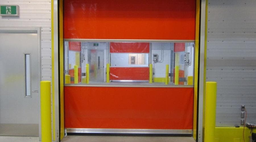 Puerta rápida industrial enrollable de PVC roja cerrada, con banda de ventanas transparentes y bolardos de protección a los lados del vano